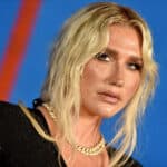 Kesha with blonde hair, wearing a black outfit and gold chain necklace against a blue background
