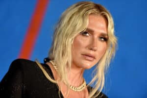 Kesha with blonde hair, wearing a black outfit and gold chain necklace against a blue background