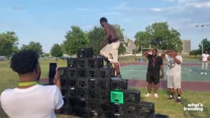 Man climbs a pyramid of stacked milk crates outdoors while others watch and record on a basketball court.