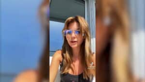 Woman with long light brown hair wearing large clear glasses and a black tank top speaking outdoors on a balcony