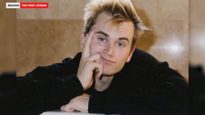 Young man with blond hair resting his head on his hand, wearing a black sweatshirt against a beige background