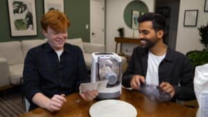 Two men smiling at a countertop dough mixer with dough inside in a modern living room setting.