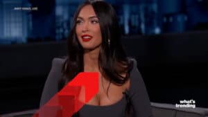 Megan Fox speaking on Jimmy Kimmel Live wearing a dark off-shoulder top with red lipstick.