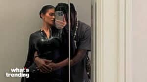 Couple taking a mirror selfie with the woman in a black shiny outfit and the man hugging her from behind.