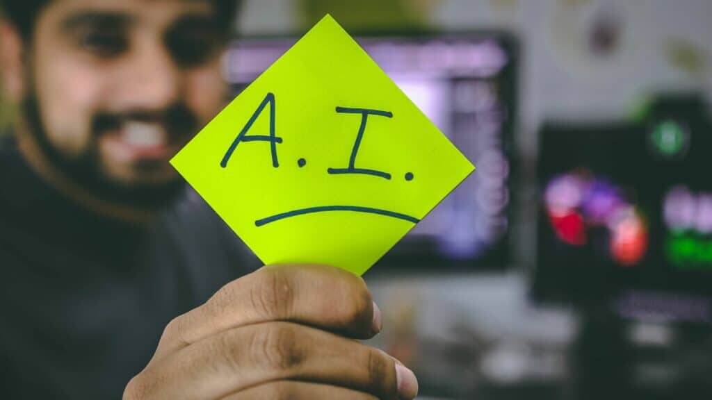Person holding a yellow sticky note with "A.I." written on it in front of a blurred computer screen background.