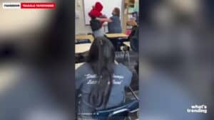 Teacher lifting a student wearing a red hoodie in a classroom with desks and seated students.