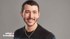 Smiling man with short curly hair, nose ring, earrings, and chain necklace wearing a black sweater against gray background