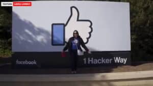 Person standing in front of Facebook's "1 Hacker Way" sign with large thumbs-up logo