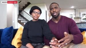 Two people sitting on a couch in a living room, one wearing glasses and a black hat, the other in a maroon sweater.