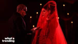 Tattooed man reading from a book to a woman in a red veil and dress under dim candlelight.