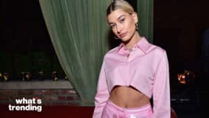 Hailey Bieber wearing a pink cropped shirt and matching pants posing indoors with green curtains in the background