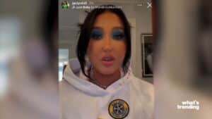 Jaclyn Hill wearing a white hoodie with a circular patch and bold blue eyeshadow speaking in a video.