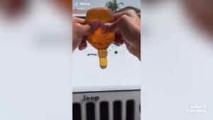 Hands squeezing an orange bottle with liquid above the front grille of a white Jeep vehicle outdoors