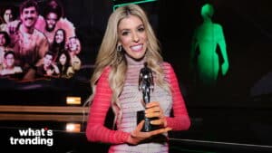 Blonde woman in pink striped dress holding a black trophy with a smiling expression at an event with "what's trending" text.