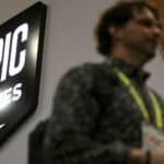 Blurred person standing near an illuminated Epic Games logo sign on a wall.