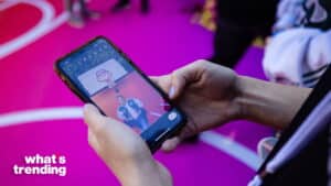 Person holding smartphone showing video of another person standing under a basketball hoop on a pink court with "what's trending" text visible