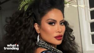 Woman with voluminous curly hair, bold makeup, and layered pearl necklace looking over her shoulder.