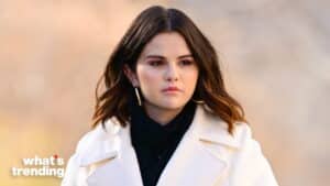 Selena Gomez wearing a white coat and black scarf with a neutral expression against a blurred background