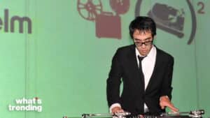 Man in black suit and glasses DJing on turntables against a green backdrop with "what's trending" text