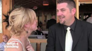 A man in a black suit and white tie smiling at a blonde woman in a white dress inside a restaurant.