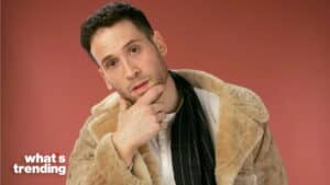 Man in beige fur coat and black scarf posing thoughtfully against a red background with "what's trending" text
