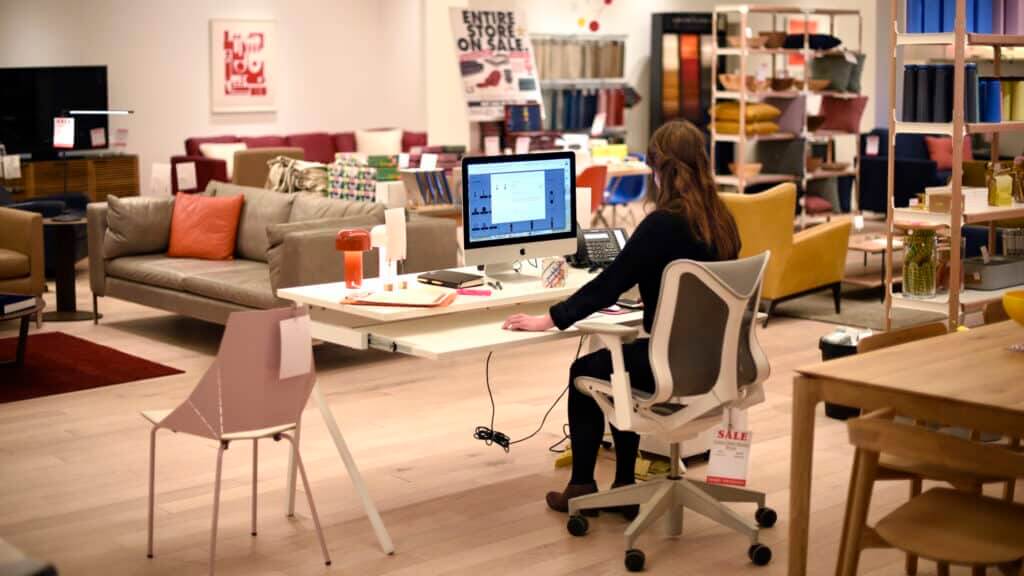 Person working on an iMac at a desk in a furniture store with sofas and chairs on sale in the background