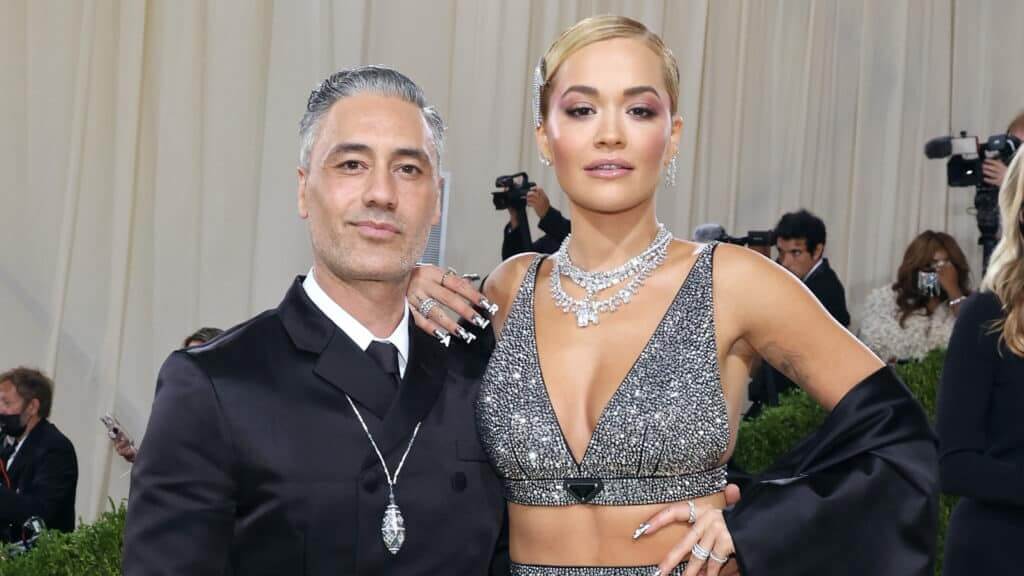 Taika Waititi and Rita Ora posing at a formal event with Rita wearing a sparkling silver Prada outfit and jewelry.