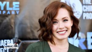 Woman with short wavy red hair smiling, wearing a green top at a branded event backdrop.