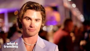 Close-up of a young man with styled hair wearing a light purple blazer and a pearl necklace at a nighttime event
