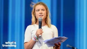 Greta Thunberg speaking into a microphone while holding papers on stage with a blue background