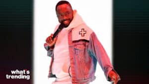 Man holding microphone wearing white hoodie and denim jacket with cross patch, "what's trending" logo visible