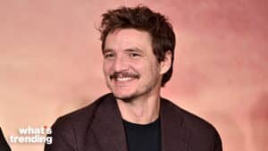 Pedro Pascal smiling wearing a dark blazer and black shirt against a pink background