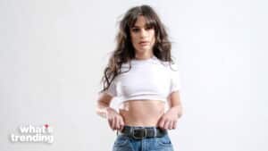 Young woman with long dark hair wearing a white cropped t-shirt and blue jeans adjusting her belt against a white background