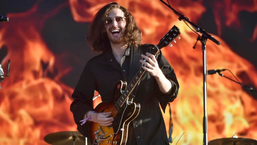 Musician with long hair and sunglasses playing a Gibson electric guitar on stage with a fiery background.