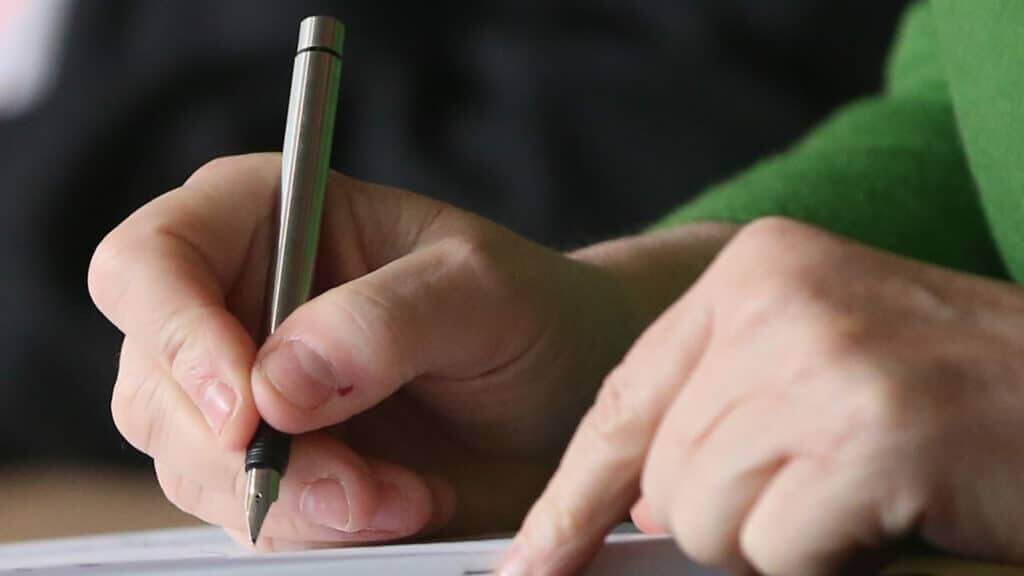 Hand holding a fountain pen poised to write on paper with the other hand pointing on the page.