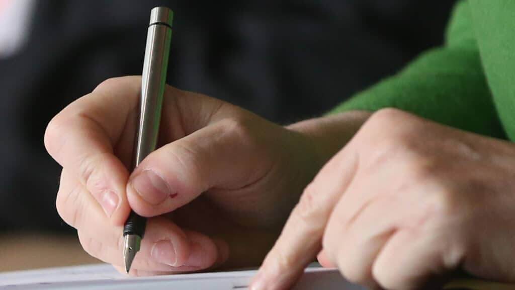 Close-up of a hand holding a fountain pen poised to write on paper with another hand pointing on the page.