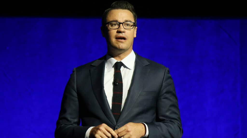 Man in glasses and dark suit speaking against a blue background