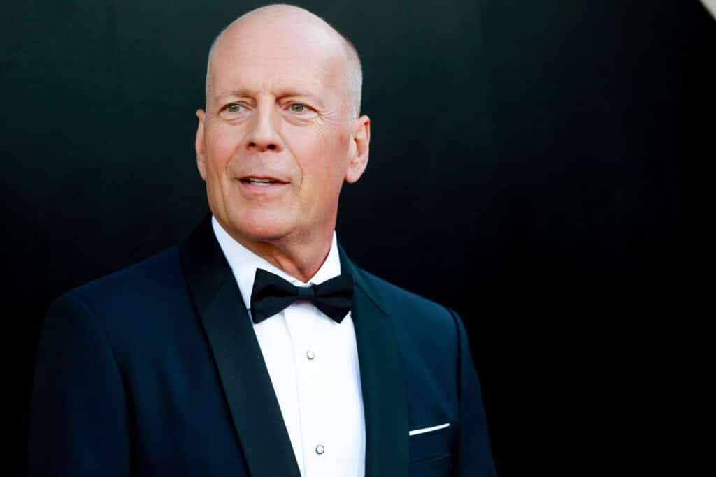 Bruce Willis wearing a black tuxedo and bow tie against a dark background