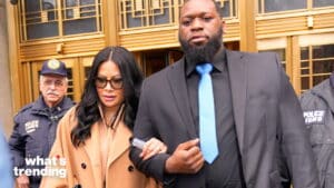 Woman in tan coat and sunglasses escorted by man in black suit and blue tie outside building with police officers behind them