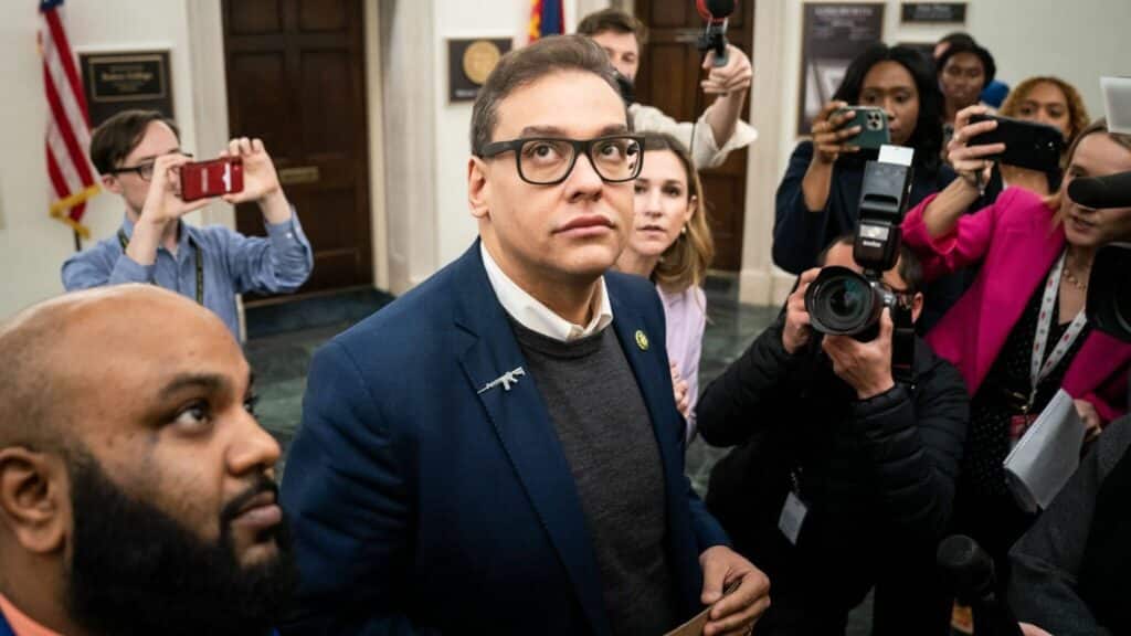 Man in glasses and blue blazer with gun-shaped pin surrounded by photographers and reporters indoors