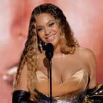 Beyoncé smiling and speaking at a microphone while holding a Grammy award, wearing a gold dress and black gloves.