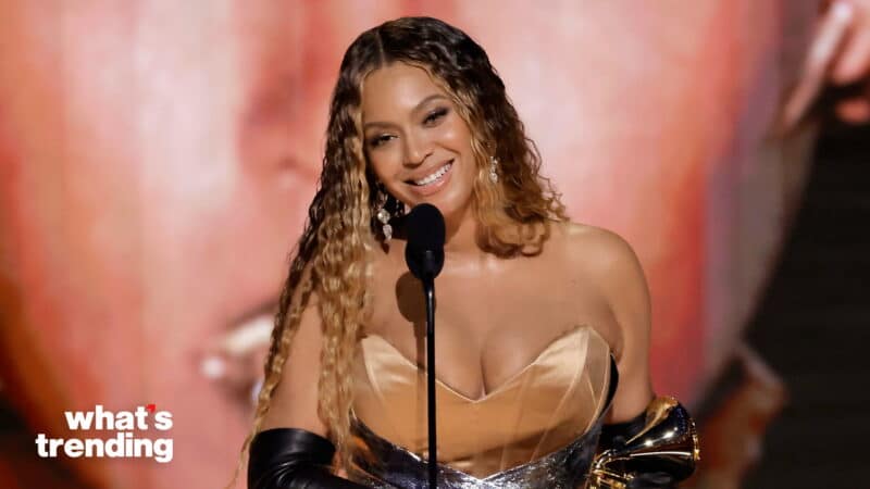 Beyoncé smiling and speaking at a microphone while holding a Grammy award, wearing a gold dress and black gloves.
