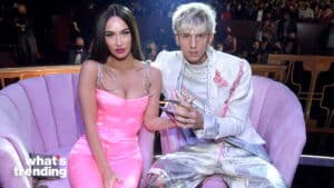 Megan Fox in a pink outfit and Machine Gun Kelly in a white suit with pink accents sitting on a lavender couch at an event.