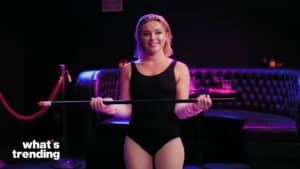 Florence Pugh in a black leotard holding a long stick in a dimly lit lounge with purple lighting