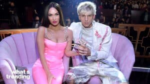 Megan Fox in a pink dress and Machine Gun Kelly in a white suit with pink accents sitting on a lavender couch at an event.
