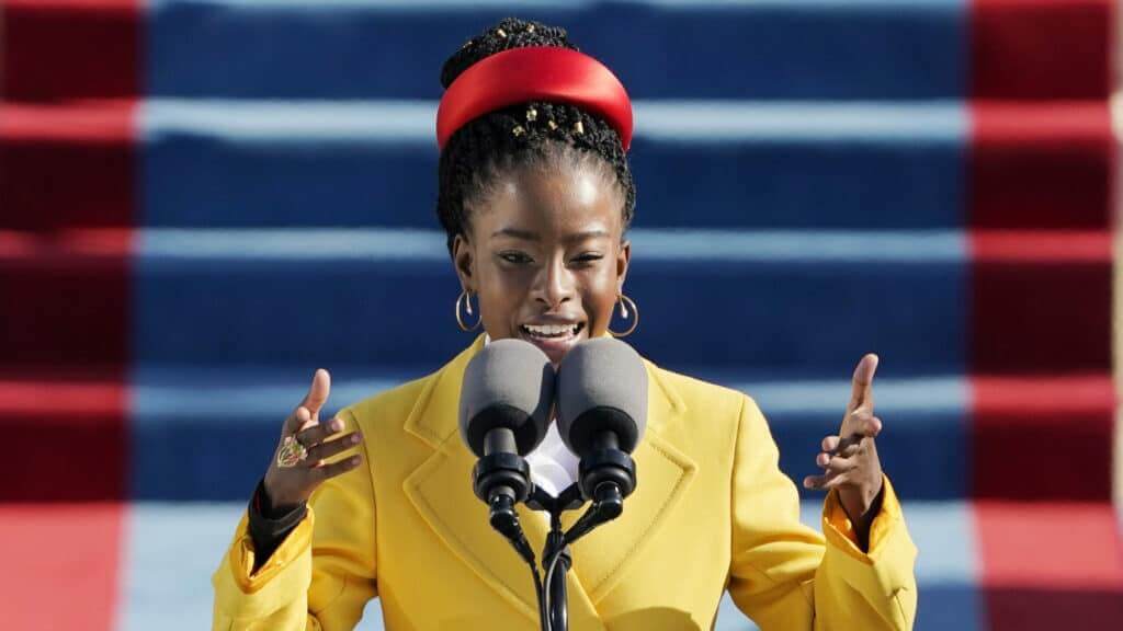 Amanda Gorman delivering a speech wearing a yellow coat and red headband at a formal event with blue and red stairs background.