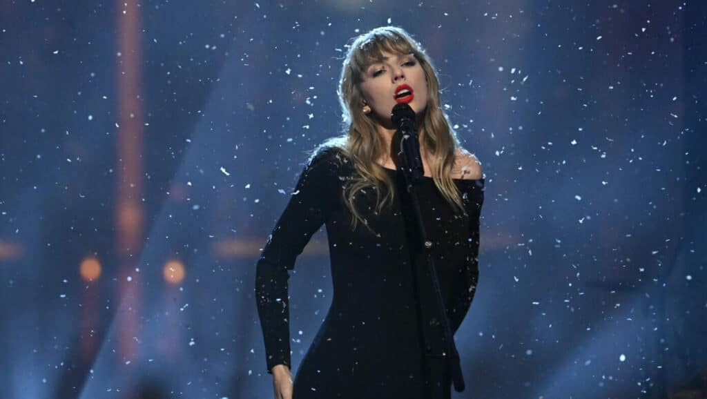 Taylor Swift singing on stage with falling snow effects, wearing a black off-shoulder dress and red lipstick