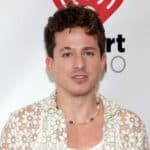 Charlie Puth wearing a white floral lace shirt and beaded necklace at an iHeartRadio event.