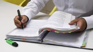 Person in white shirt writing notes in a thick document with colorful tabs and a green Stabilo Boss highlighter nearby