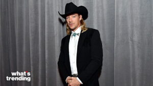 Man in black cowboy hat, black jacket, white shirt, and turquoise bolo tie standing against gray curtain background